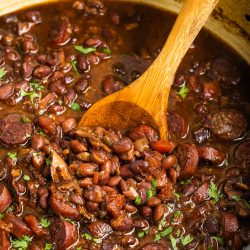 A pot of cooked red beans mixed with slices of sausage, garnished with chopped herbs, with a wooden spoon stirring the dish.