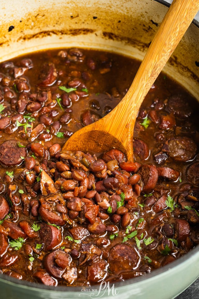 A pot of cooked red beans mixed with slices of sausage, garnished with chopped herbs, with a wooden spoon stirring the dish.