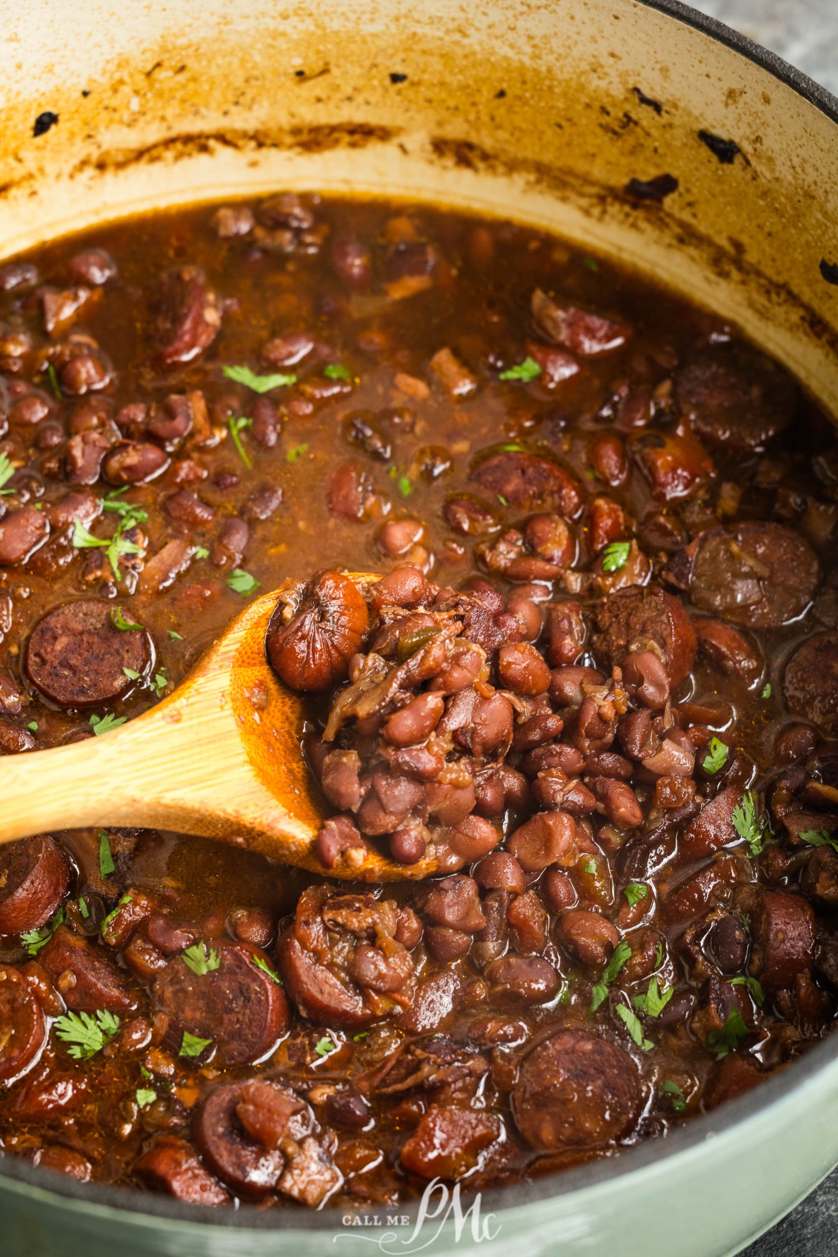 A pot of homemade red beans and sausage stew with a wooden spoon stirring the thick, seasoned mixture.