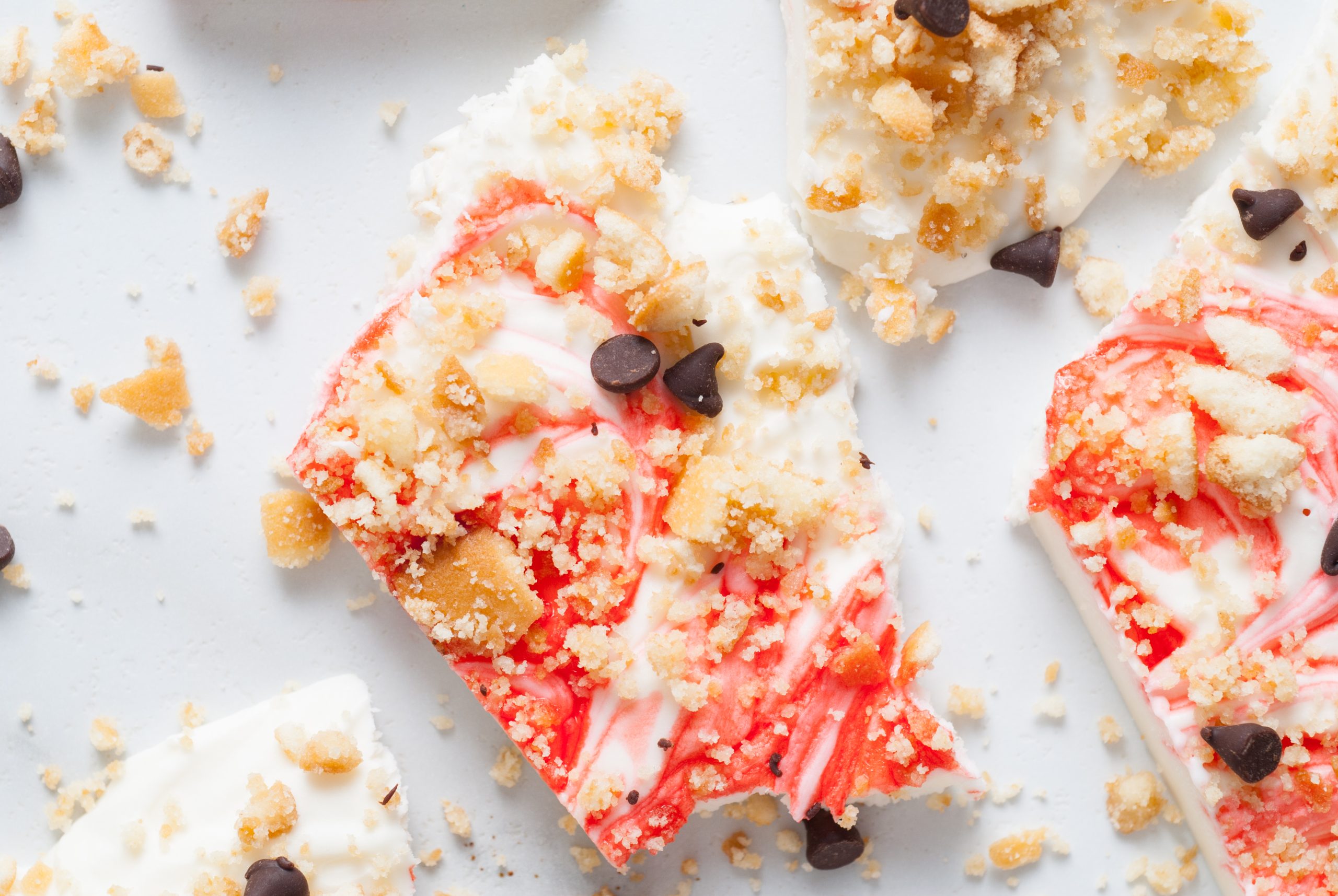 Close-up of broken pieces of white chocolate bark with red swirls, crumbly cookie pieces, and chocolate chips scattered on a white surface.