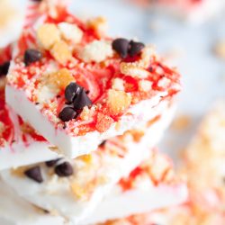 Close-up of a stack of strawberry yogurt bark topped with granola and chocolate chips on a white surface. The yogurt has a marbled red and white appearance.