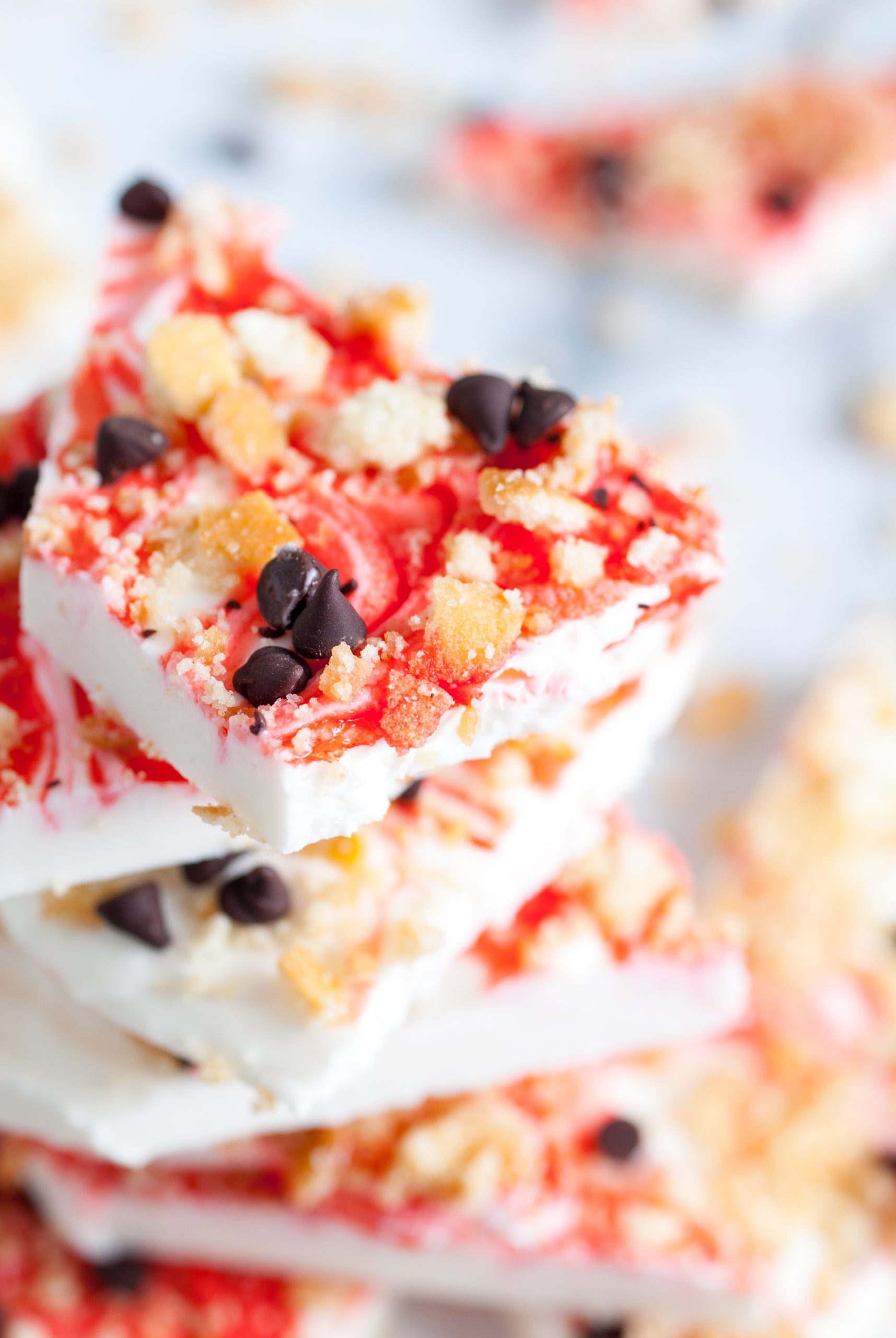 Close-up of a stack of strawberry yogurt bark topped with granola and chocolate chips on a white surface. The yogurt has a marbled red and white appearance.