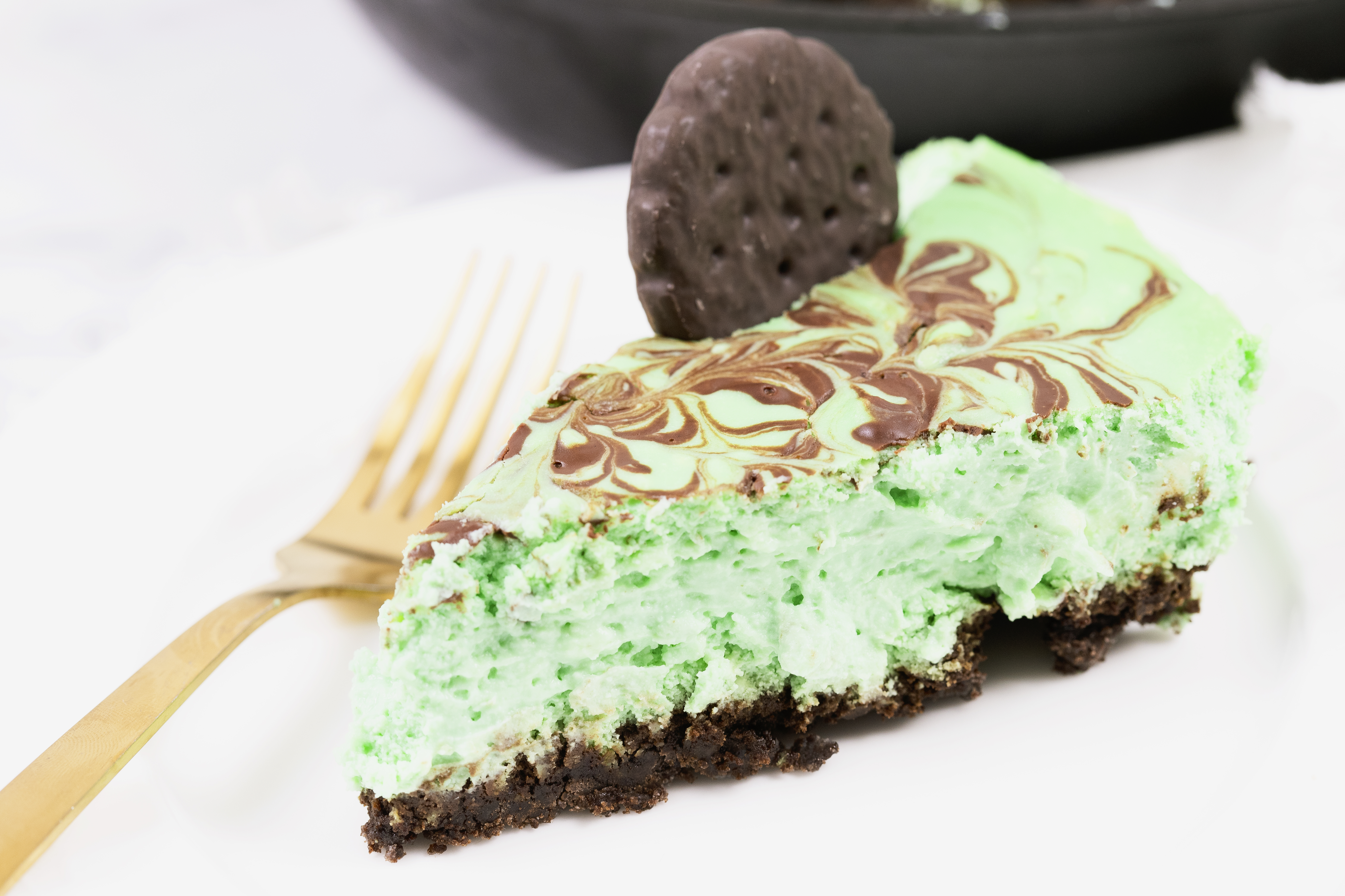 A slice of green mint chocolate dessert on a plate with a fork, topped with a chocolate cookie, with the remaining dessert visible in the background.