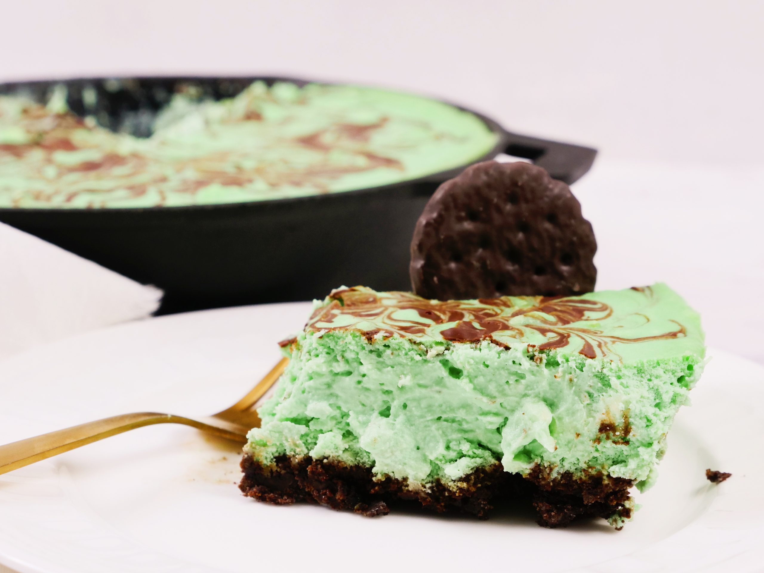 A slice of green mint chocolate dessert on a plate with a fork, topped with a chocolate cookie, with the remaining dessert visible in the background.