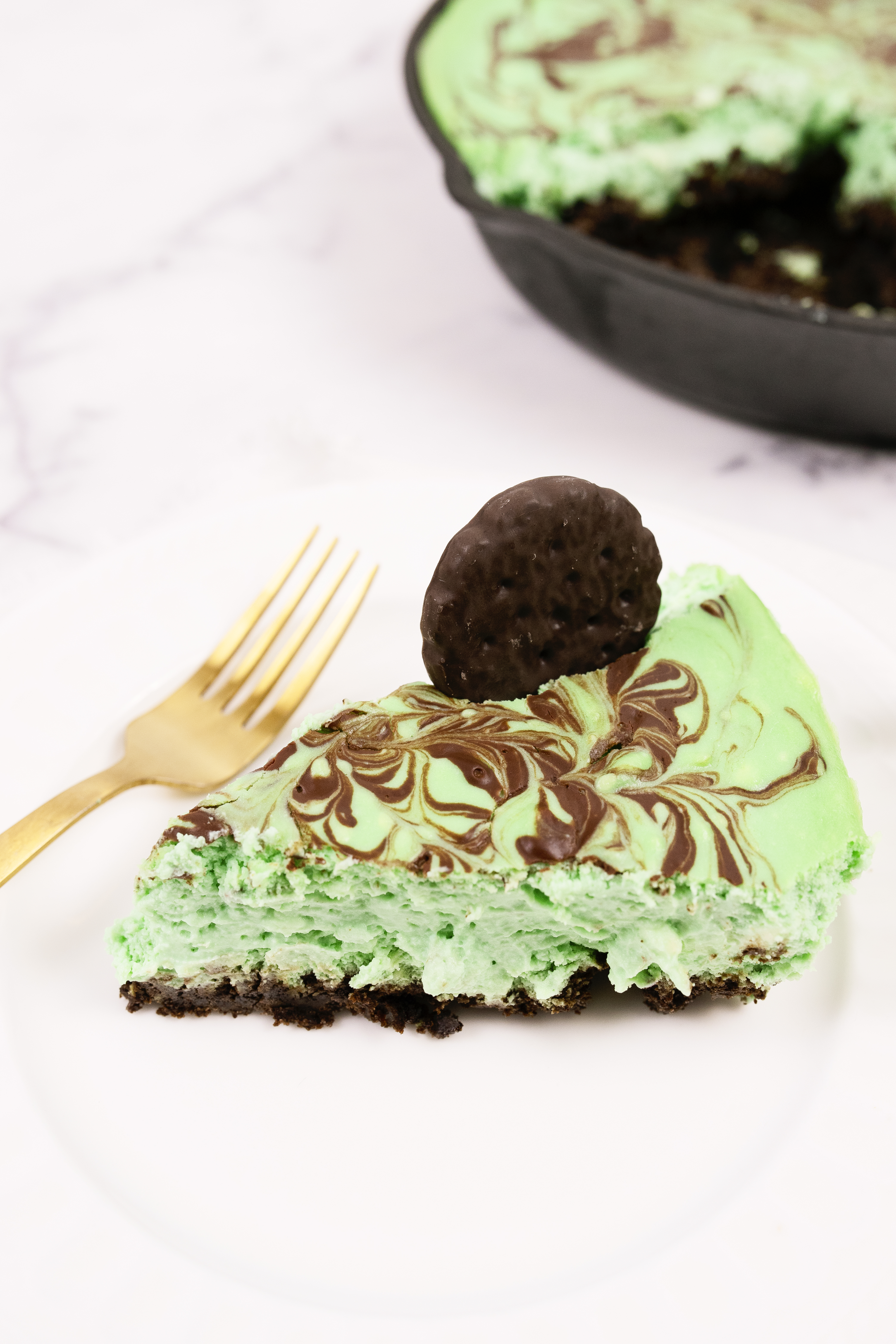 A slice of green mint chocolate dessert on a plate with a fork, topped with a chocolate cookie, with the remaining dessert visible in the background.