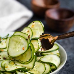 A bowl of sliced cucumber salad being lifted by a fork, with two wooden bowls in the background.