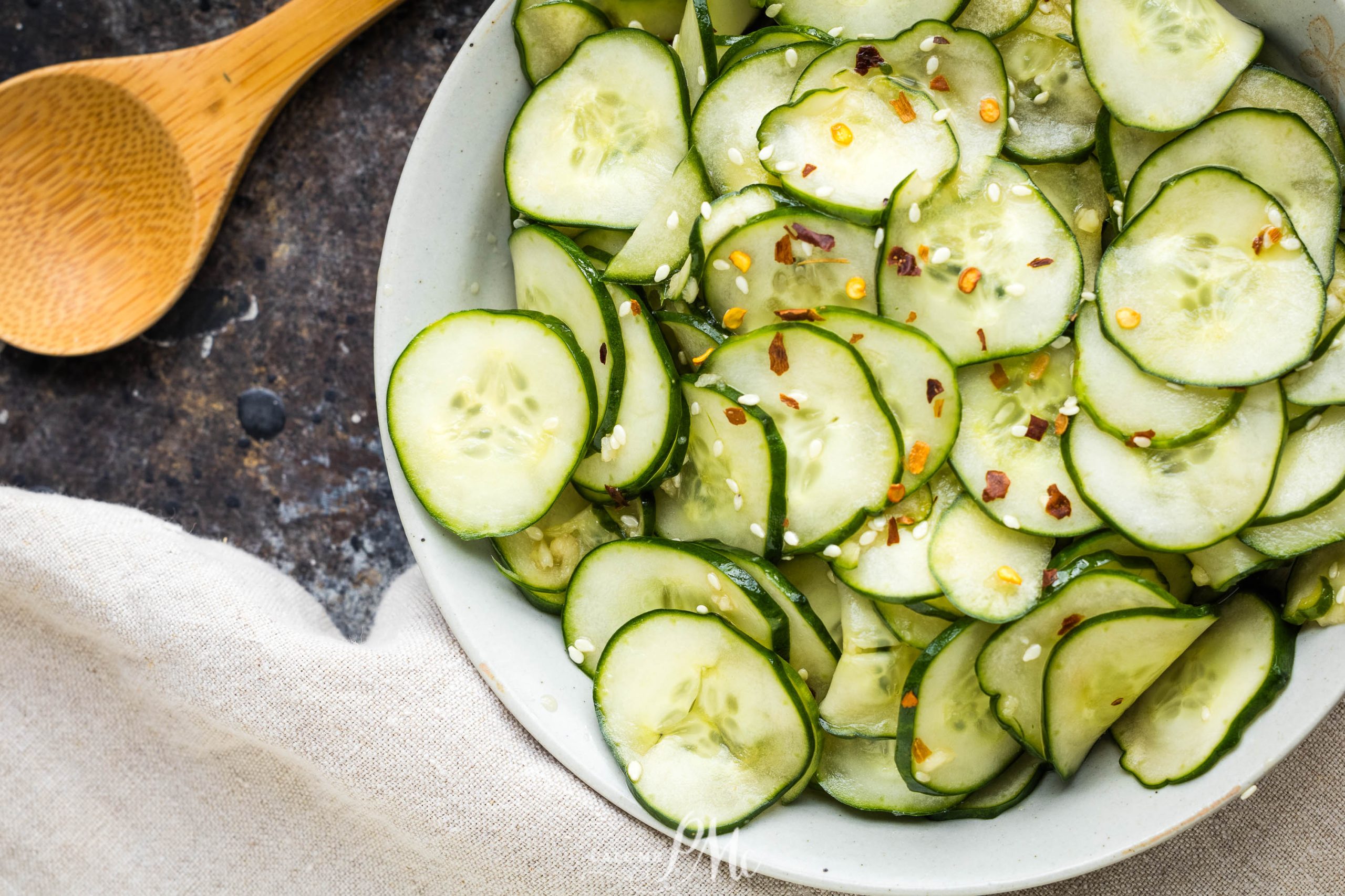 A bowl of sliced cucumbers seasoned with red pepper flakes and sesame seeds sits on a dark surface next to a wooden spoon and a beige cloth.