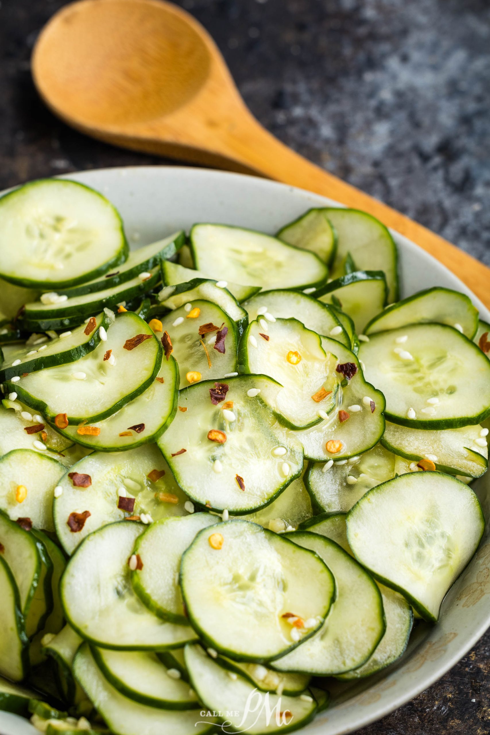 A bowl of sliced cucumbers topped with red pepper flakes and herbs, with a wooden spoon in the background.