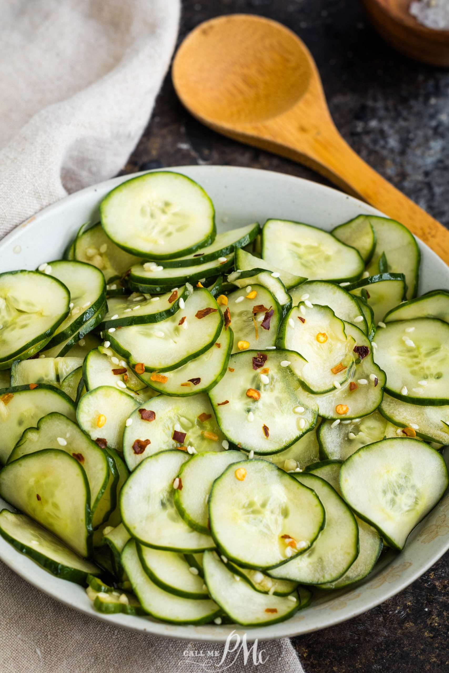 A bowl of cucumber salad, garnished with red pepper flakes and sesame seeds, with a wooden spoon laid next to it.