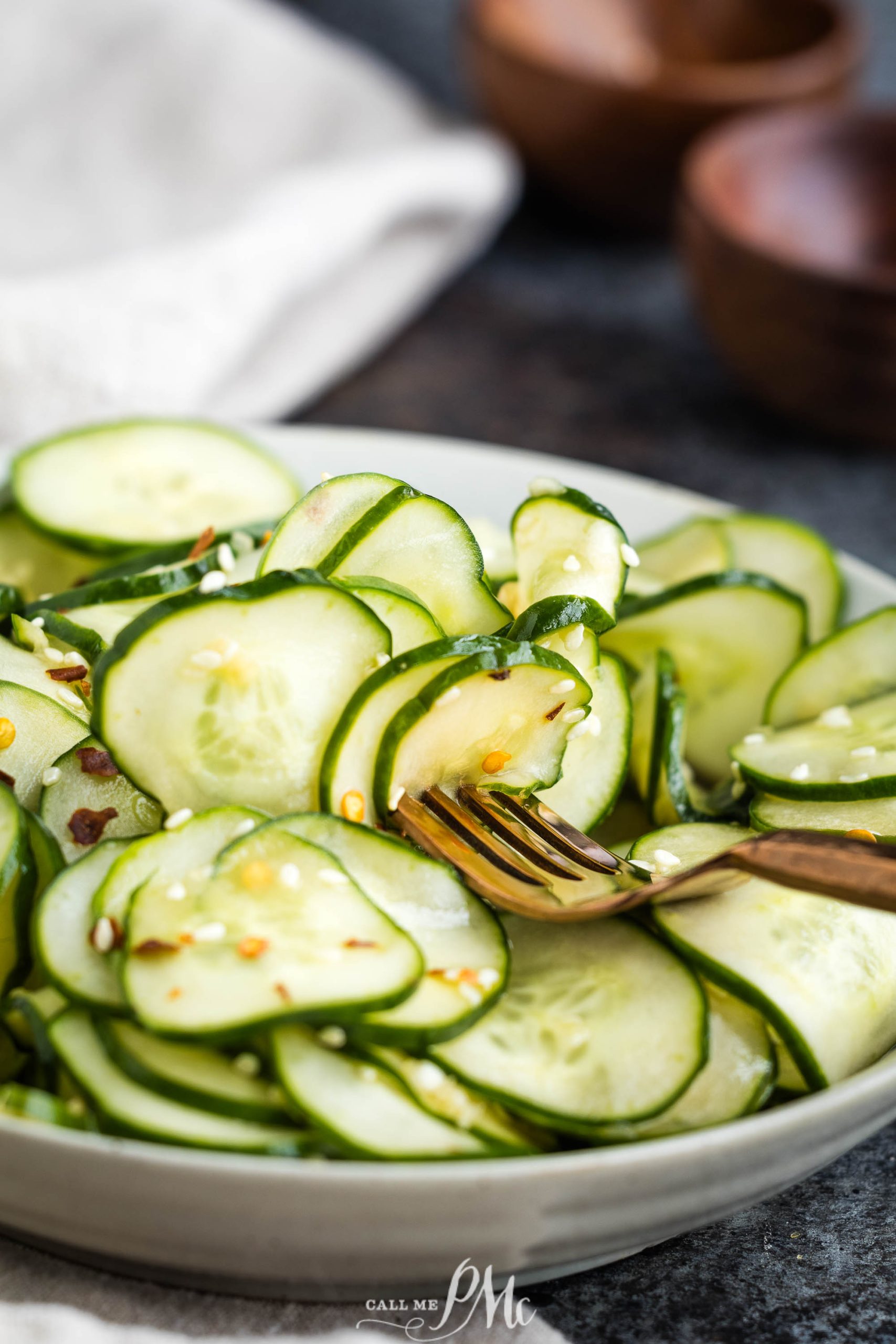 A plate of sliced cucumbers garnished with sesame seeds and chili flakes, with a fork picking up a slice.