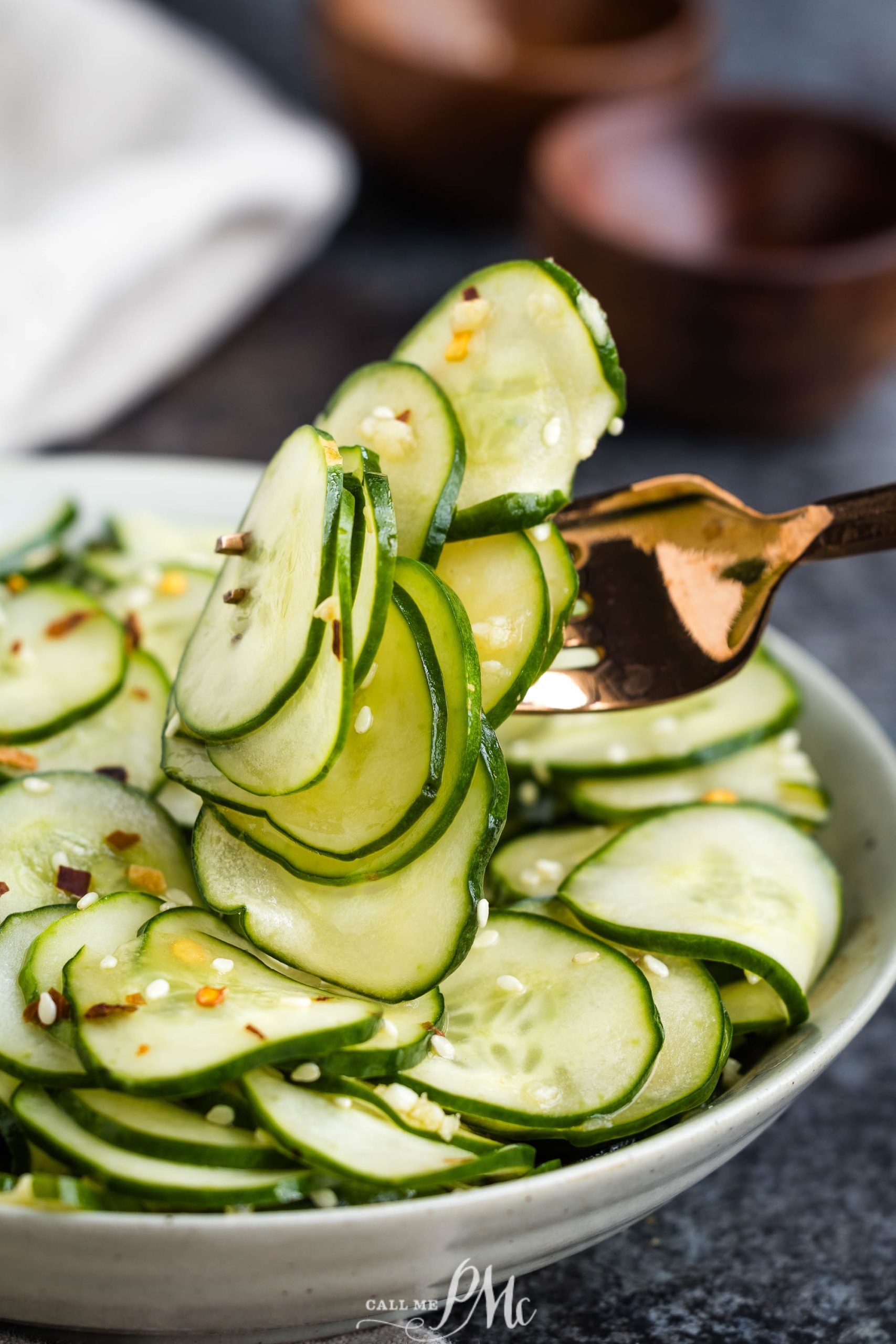 A fork lifts a slice of cucumber from a bowl filled with seasoned, thinly sliced cucumber. Two wooden bowls and a napkin are visible in the background.