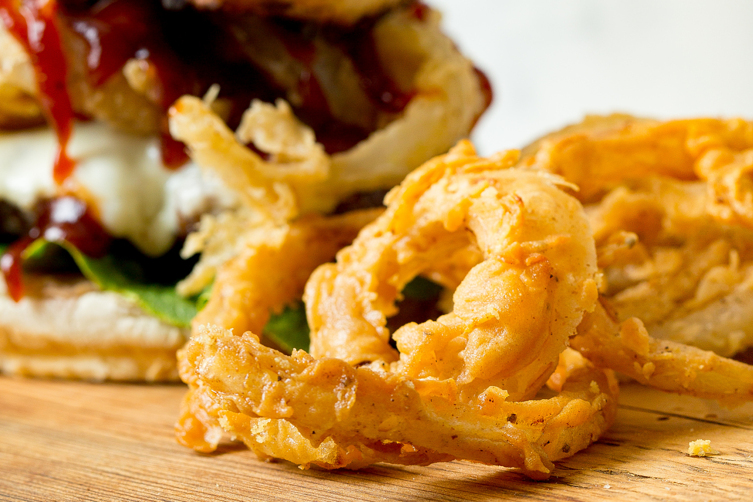 Close-up of crispy onion rings served on a wooden surface with a sandwich topped with sauce and lettuce in the background.