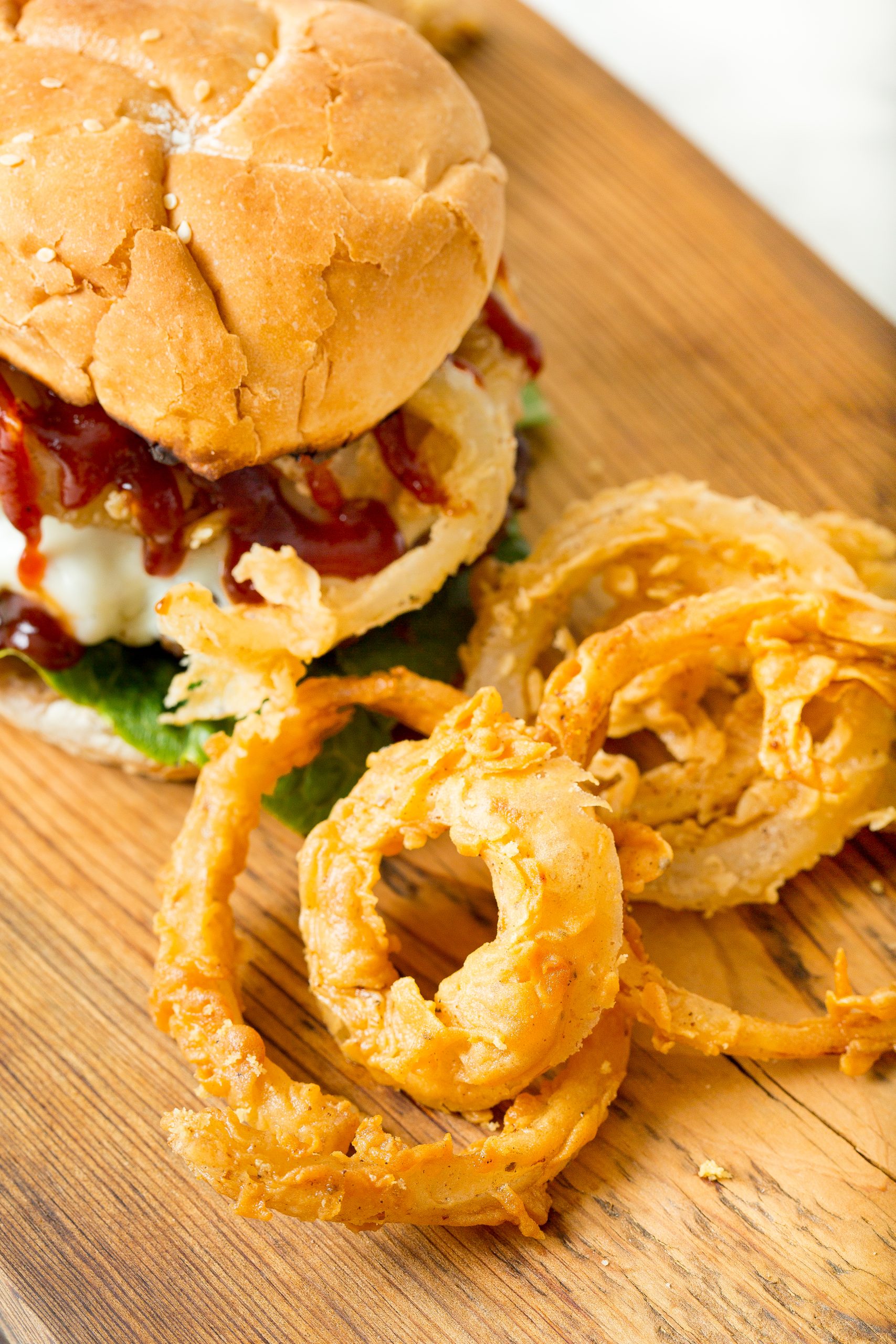 Close-up of a burger topped with sauce and vegetables on a wooden board, accompanied by crispy onion rings.