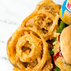 Close-up of crispy golden onion rings, a bottle of sauce, and part of a burger with lettuce, tomato, and cheese on a wooden board.