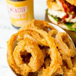 A bowl of golden, crispy onion rings, a partially visible hamburger with toppings, and a bottle of beer on a white surface.