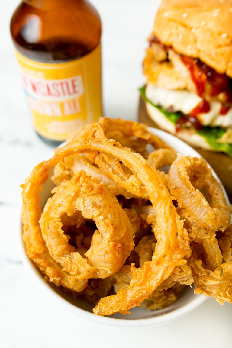 A bowl of golden, crispy onion rings, a partially visible hamburger with toppings, and a bottle of beer on a white surface.