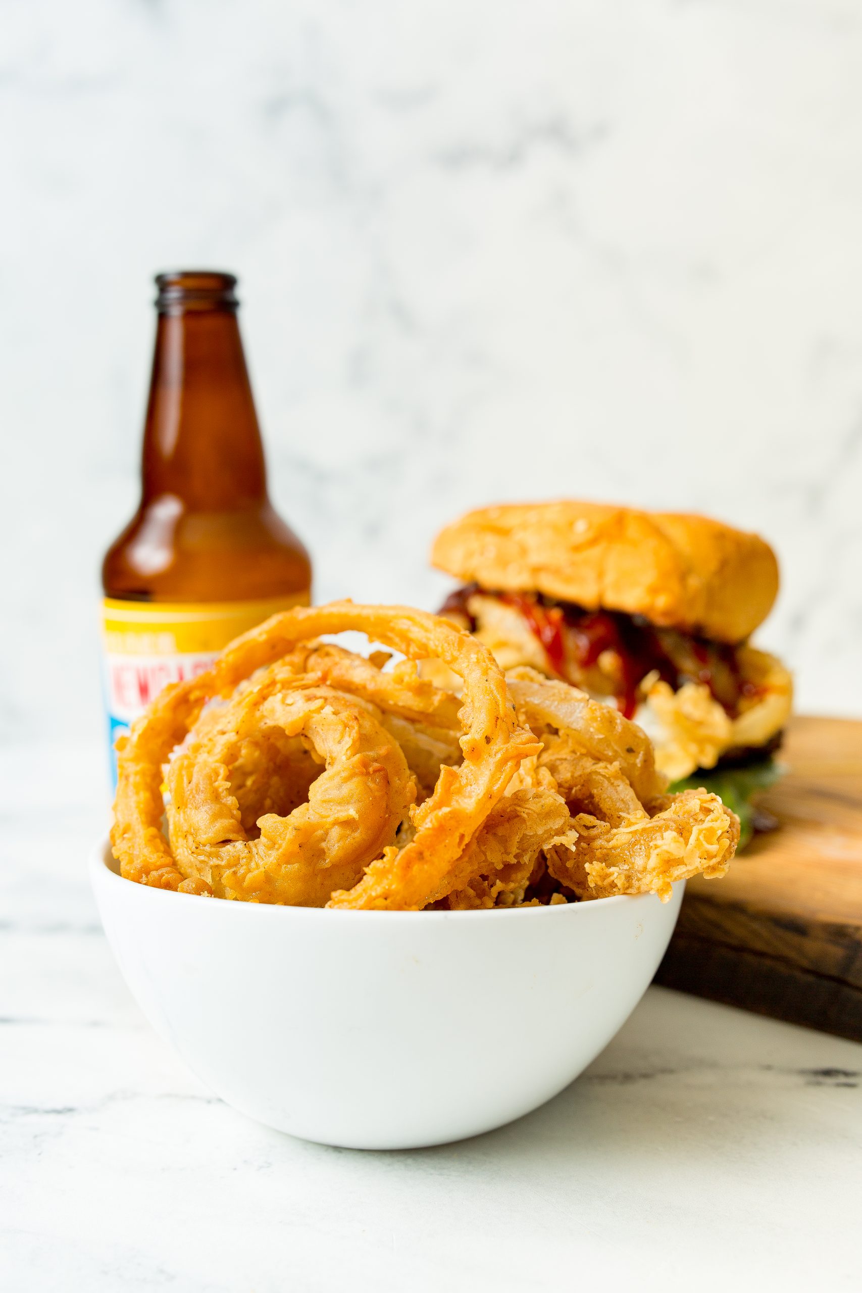 A bowl of crispy onion rings is in the foreground, with a burger and a bottle of beer in the blurred background.