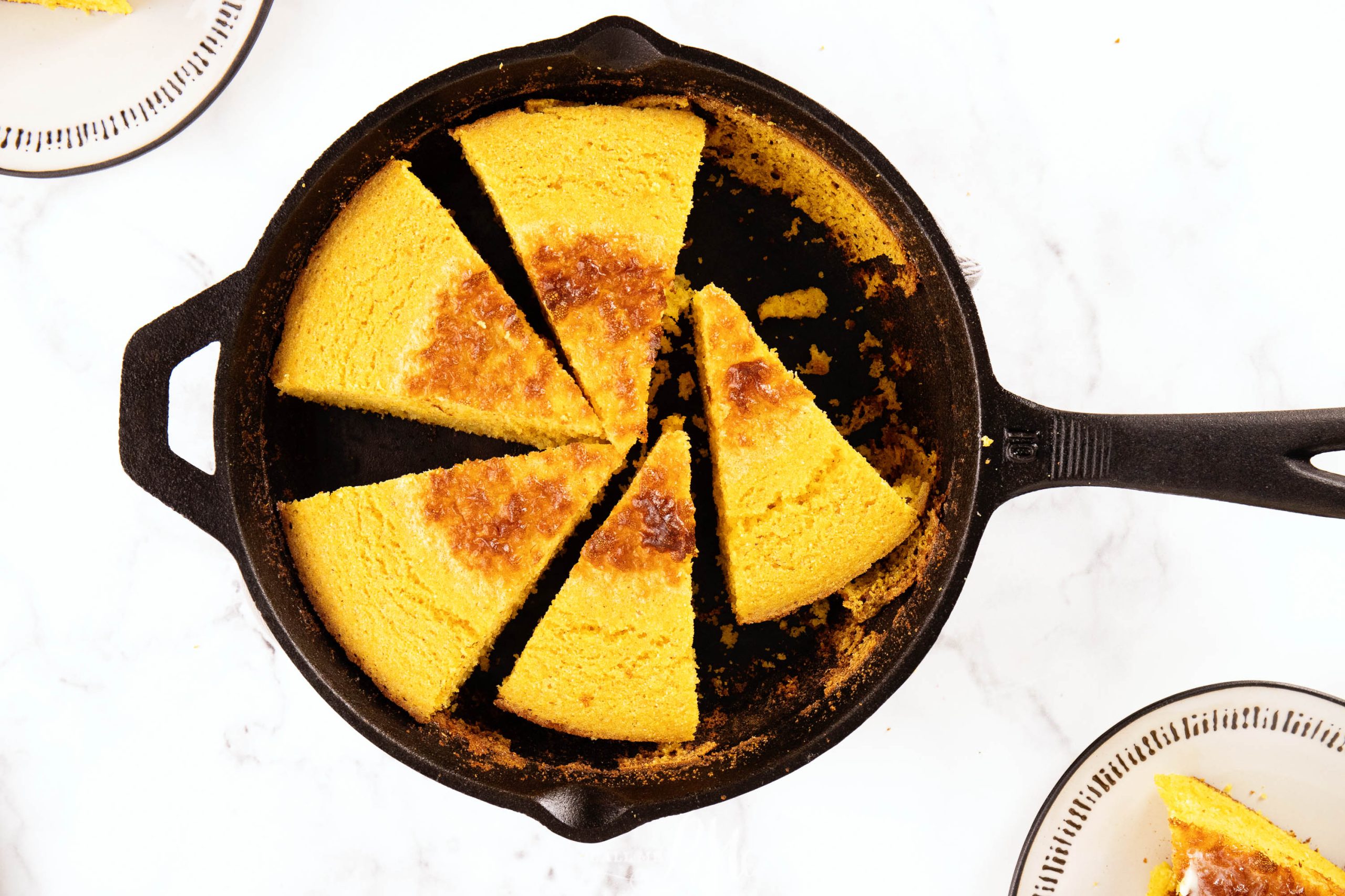 A skillet with eight triangular slices of bread, featuring a golden-brown crust on a white surface. Two plates with cornbread slices are partially visible.