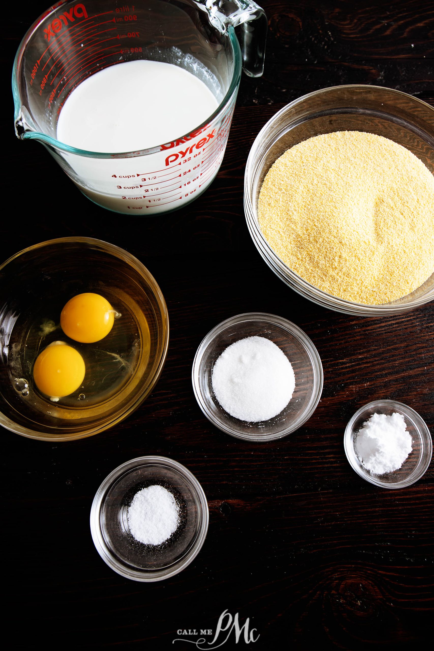 Ingredients for a recipe are laid out on a dark wooden surface, including milk in a measuring cup, two eggs in a bowl, a bowl of cornmeal, and smaller bowls of salt, sugar, and baking powder.