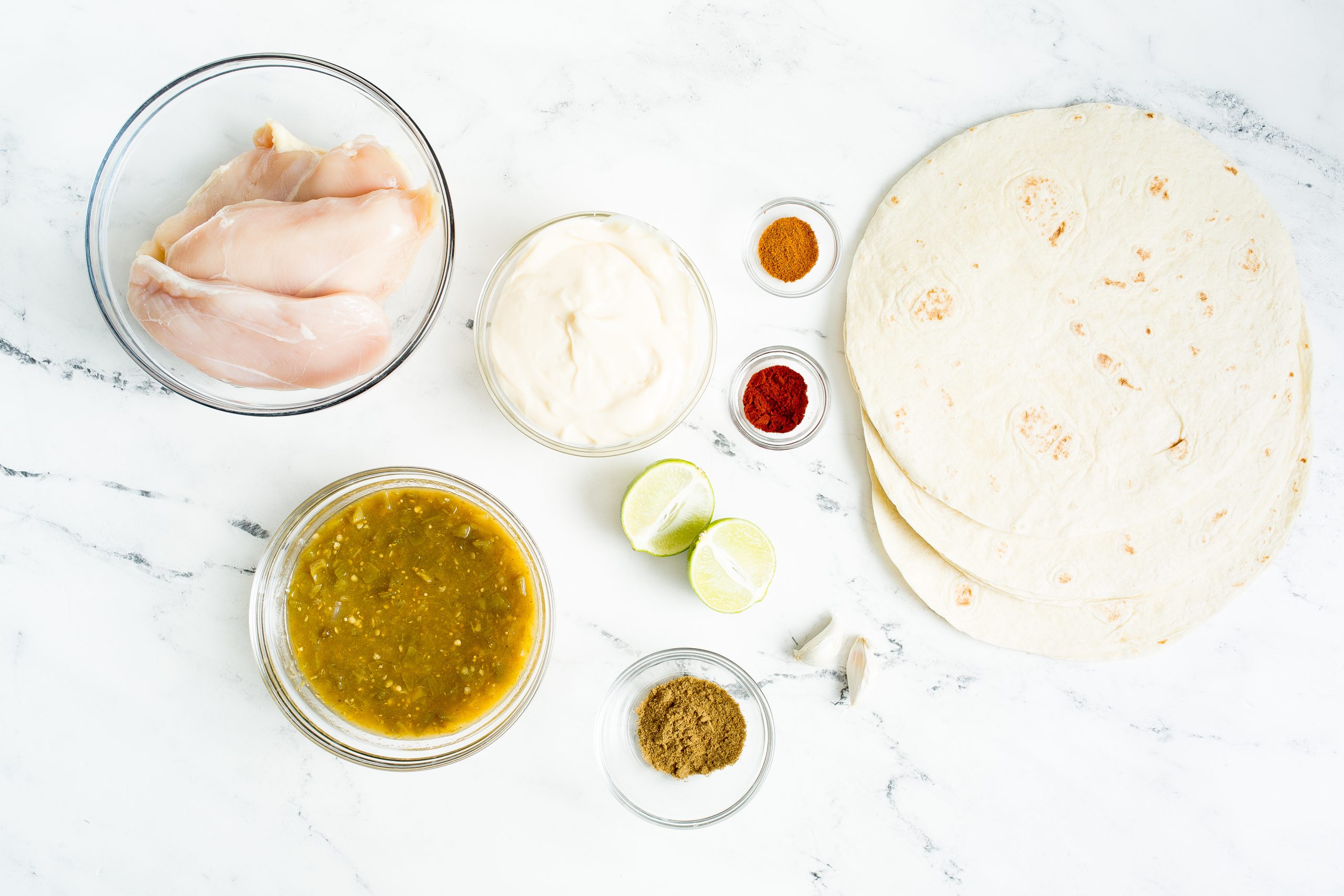 Top-down view of ingredients including raw chicken breasts, tortillas, lime halves, seasonings, grated cheese, green salsa, and mayonnaise arranged on a white surface.