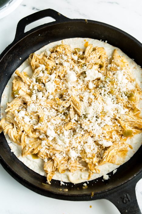 A cast iron skillet containing a tortilla topped with shredded chicken, green chili, and crumbled cheese.