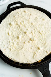 A round cast iron skillet filled with an unbaked, creamy white mixture topped with grated cheese.