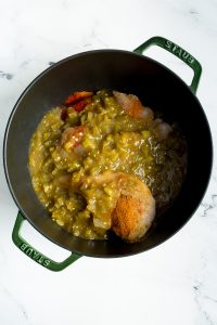 A pot containing raw chicken, chopped green chilies, and assorted spices on a marble counter.