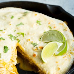 Close-up of a skillet with creamy enchiladas topped with melted cheese, garnished with cilantro and lime wedges. A portion has been cut out, revealing the filling inside.