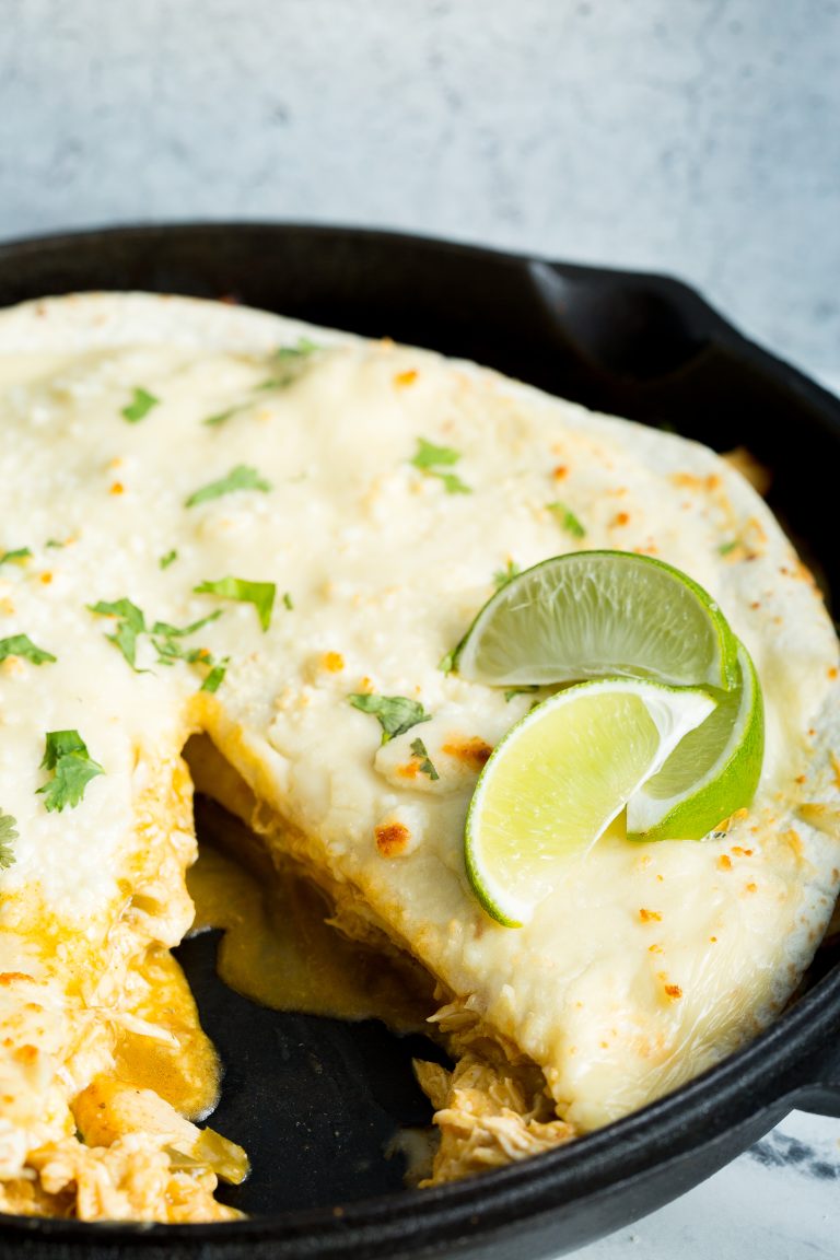 Close-up of a skillet with creamy enchiladas topped with melted cheese, garnished with cilantro and lime wedges. A portion has been cut out, revealing the filling inside.