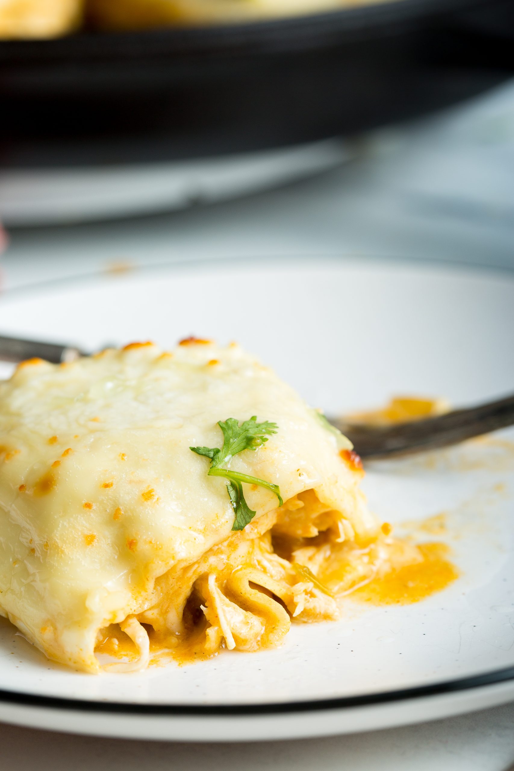 Close-up of a piece of cheesy lasagna on a white plate, garnished with a sprig of cilantro. A fork rests beside the partially eaten lasagna, revealing layers of pasta and sauce.