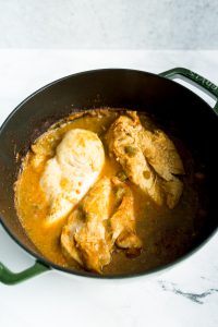 A black pot containing cooked chicken breasts in a thick, golden-brown sauce, placed on a white marble surface.