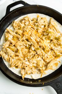 A cast-iron skillet containing shredded chicken on top of a tortilla with some sauce and green peppers.