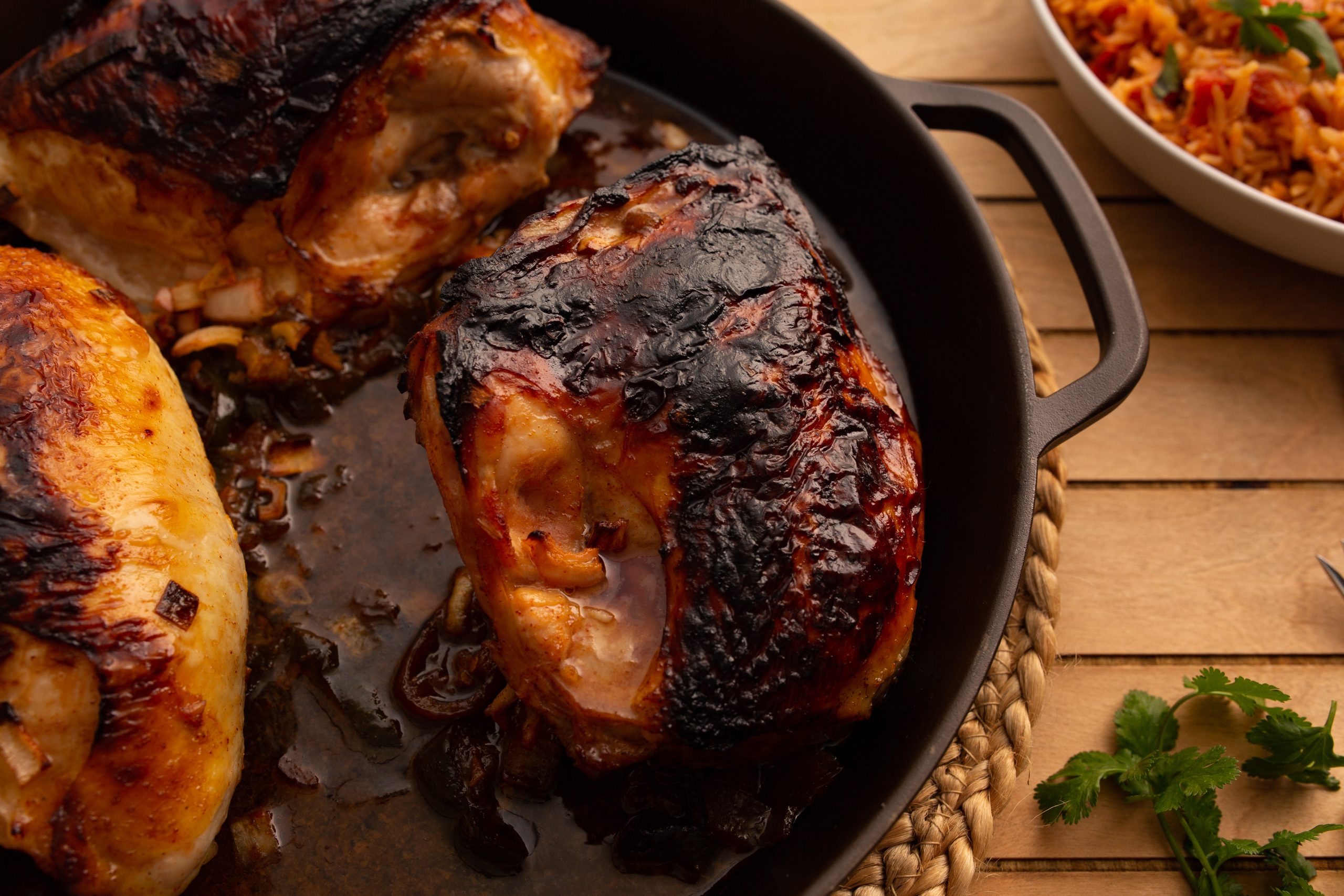 Close-up of a charred, roasted chicken thigh in a pan beside a bowl of mixed vegetables on a woven mat over a wooden table. Silverware and fresh cilantro are visible in the background.