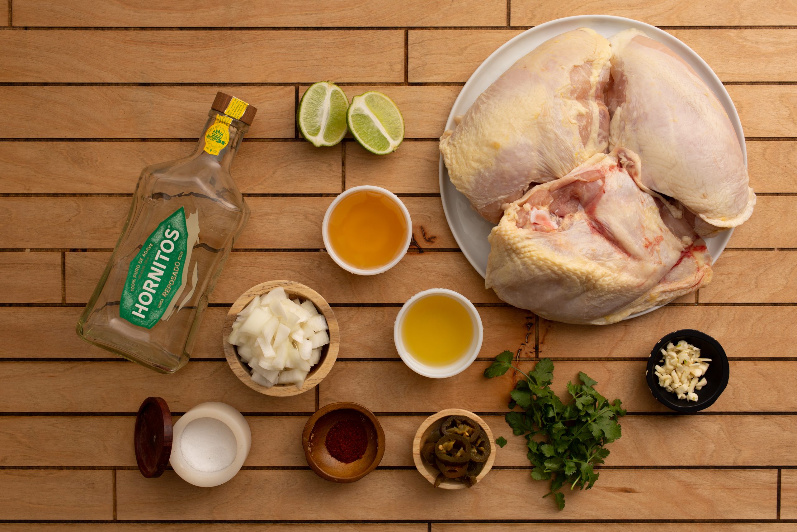 A raw whole chicken on a plate, surrounded by cooking ingredients including lime halves, tequila, diced onions, garlic, cilantro, spices, and small bowls of oil and sauce, reminiscent of a Chipotle Lime Split Breasts recipe, all arranged on a wooden surface.