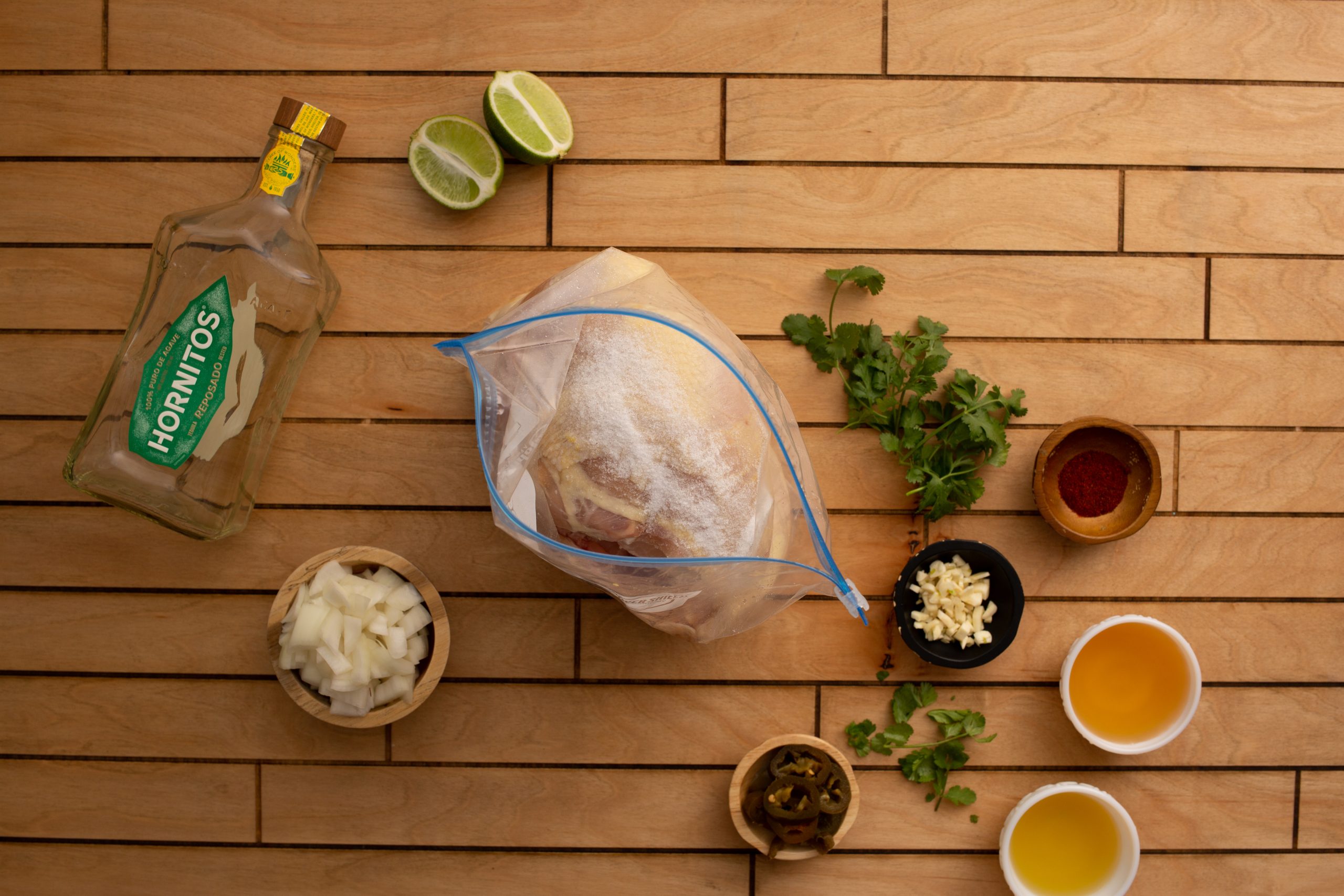 Overhead view of a wooden surface with tequila, lime wedges, diced onions, garlic, chopped cilantro, olive oil, and spices arranged around a plastic bag containing meat.