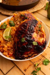 A white plate with grilled chicken, charred on the skin, alongside a serving of orange rice. Garnished with fresh cilantro and a lime wedge, set on a wooden tabletop with a pan partially visible.