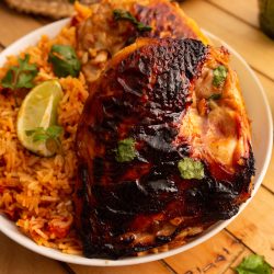 A white plate with grilled chicken, charred on the skin, alongside a serving of orange rice. Garnished with fresh cilantro and a lime wedge, set on a wooden tabletop with a pan partially visible.