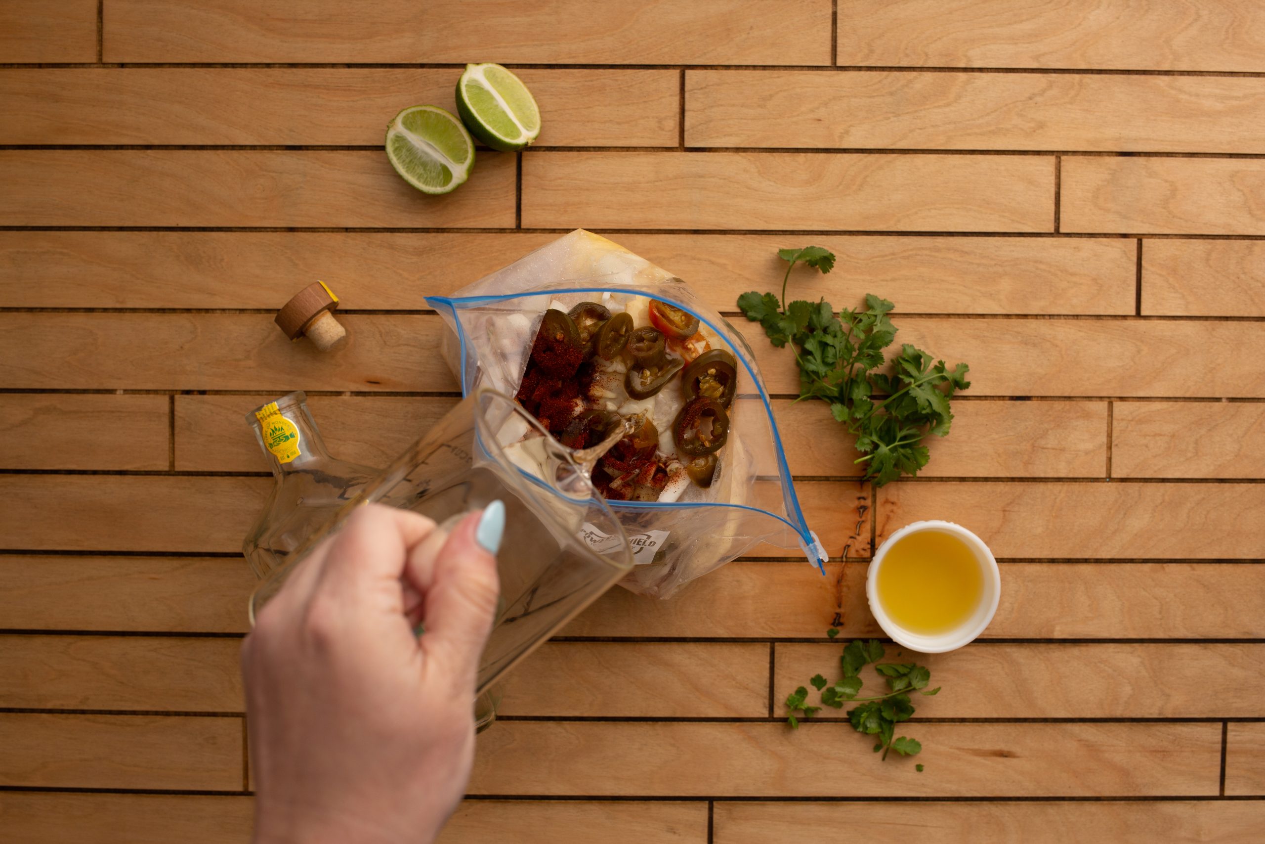 A hand pours liquid from a clear bottle into a plastic bag containing food ingredients. Lime halves, cilantro, and a small bowl of oil are on a wooden surface nearby, ready to add zest to your chipotle lime split breasts.