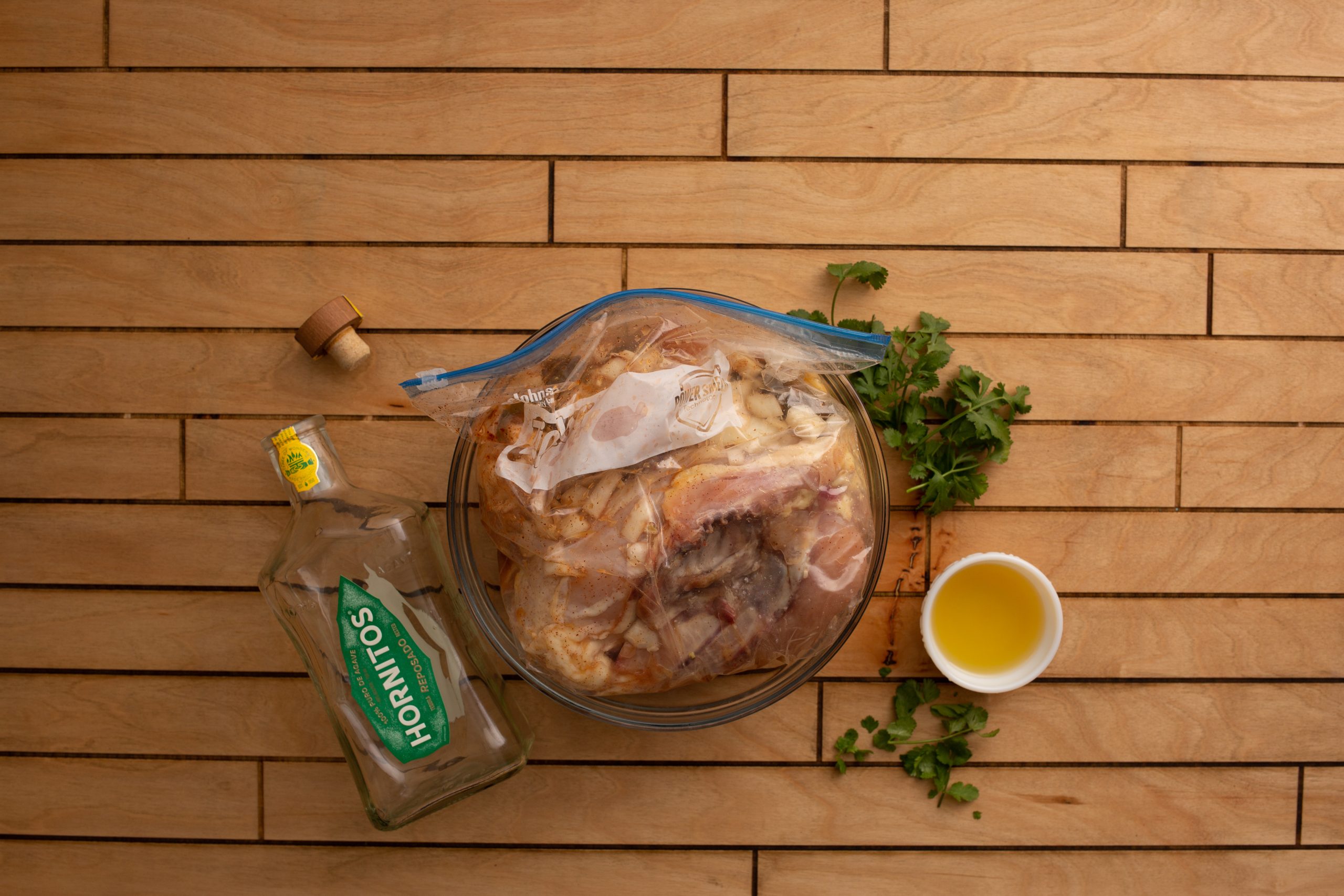 Overhead view of a wooden surface with tequila, lime wedges, diced onions, garlic, chopped cilantro, olive oil, and spices arranged around a plastic bag containing meat.