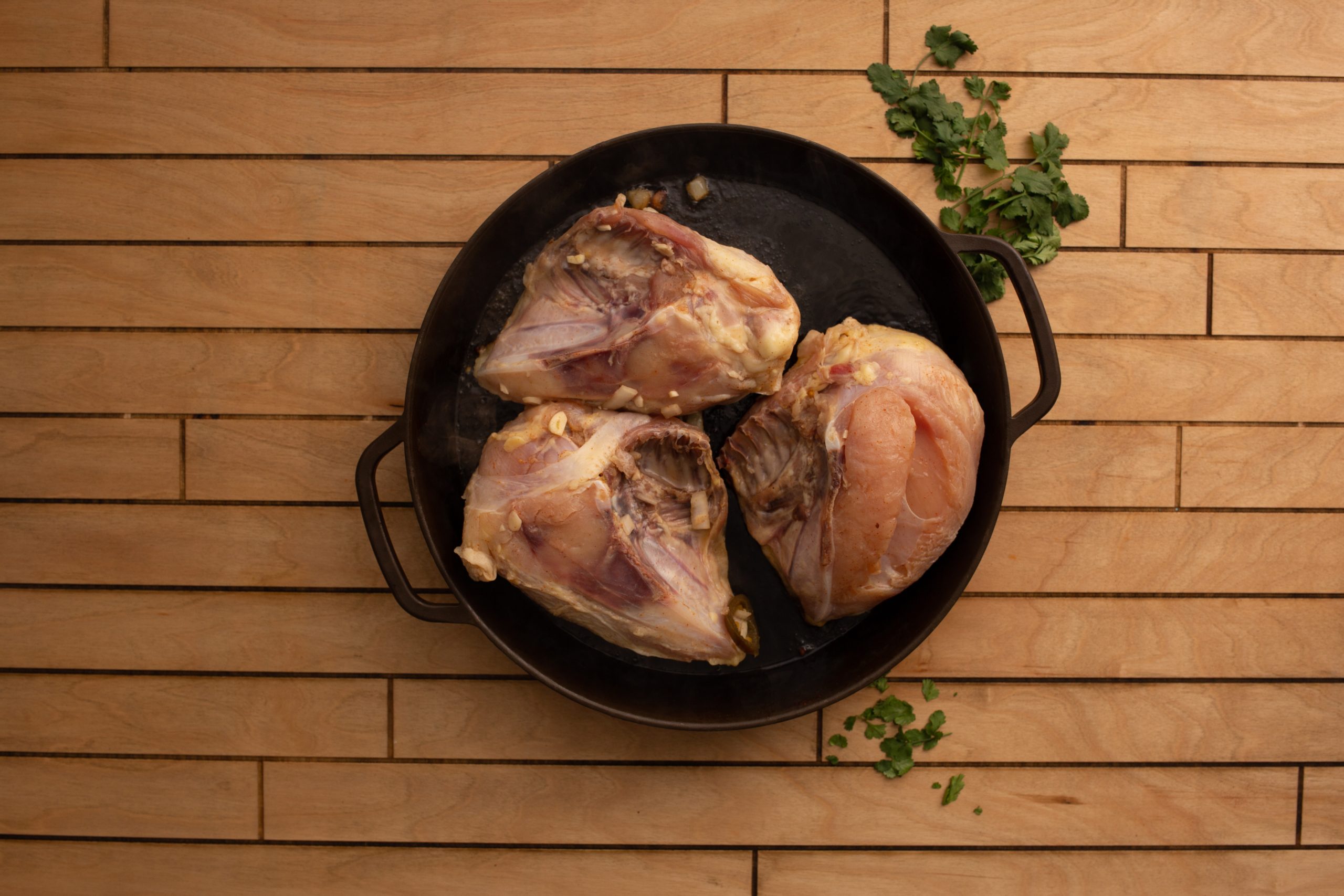 Overhead view of a wooden surface with tequila, lime wedges, diced onions, garlic, chopped cilantro, olive oil, and spices arranged around a plastic bag containing meat.