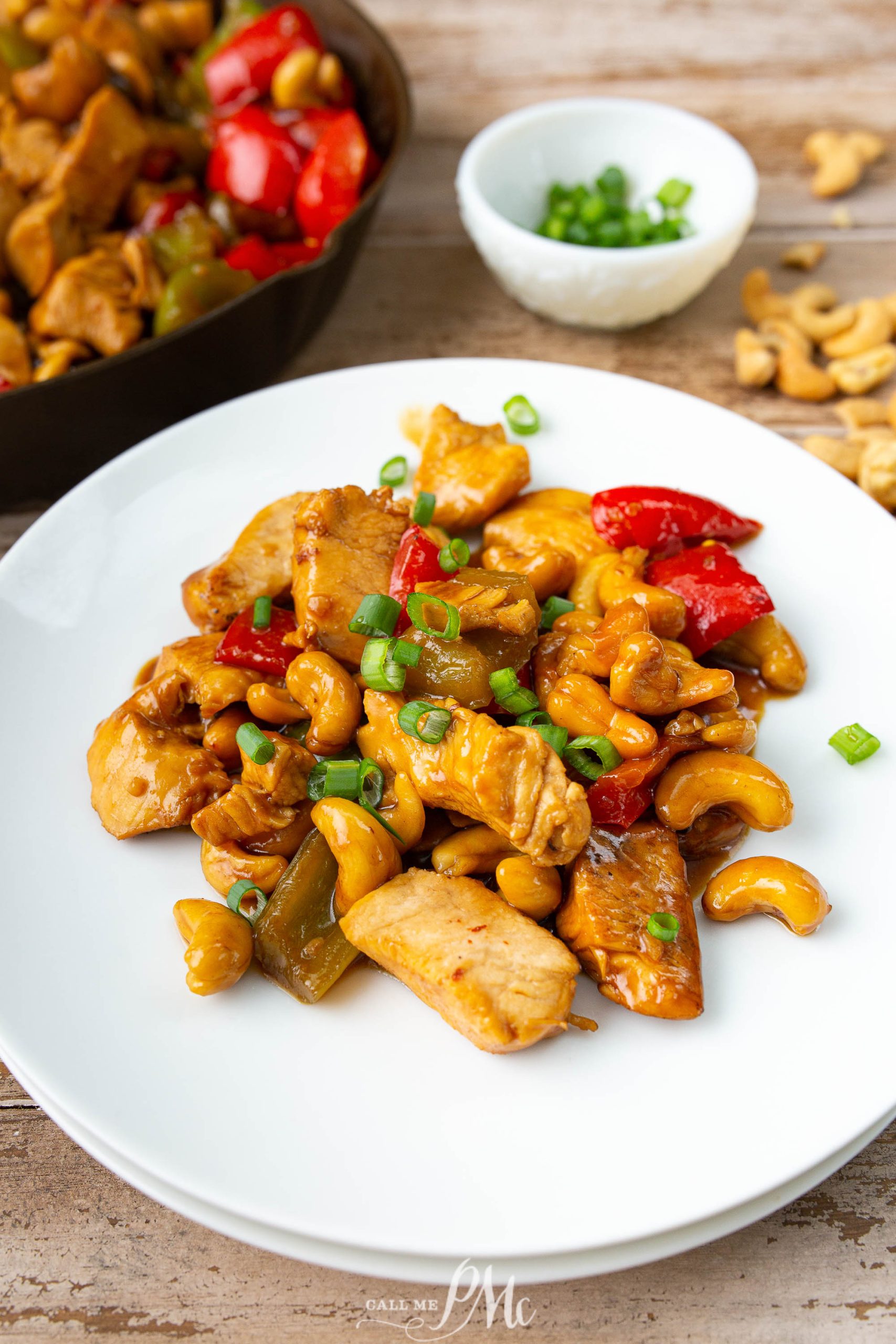 A plate of cashew chicken with bell peppers, green onions, and cashews, garnished with chopped green onions. A skillet with more of the dish and a small bowl of chopped green onions are in the background.