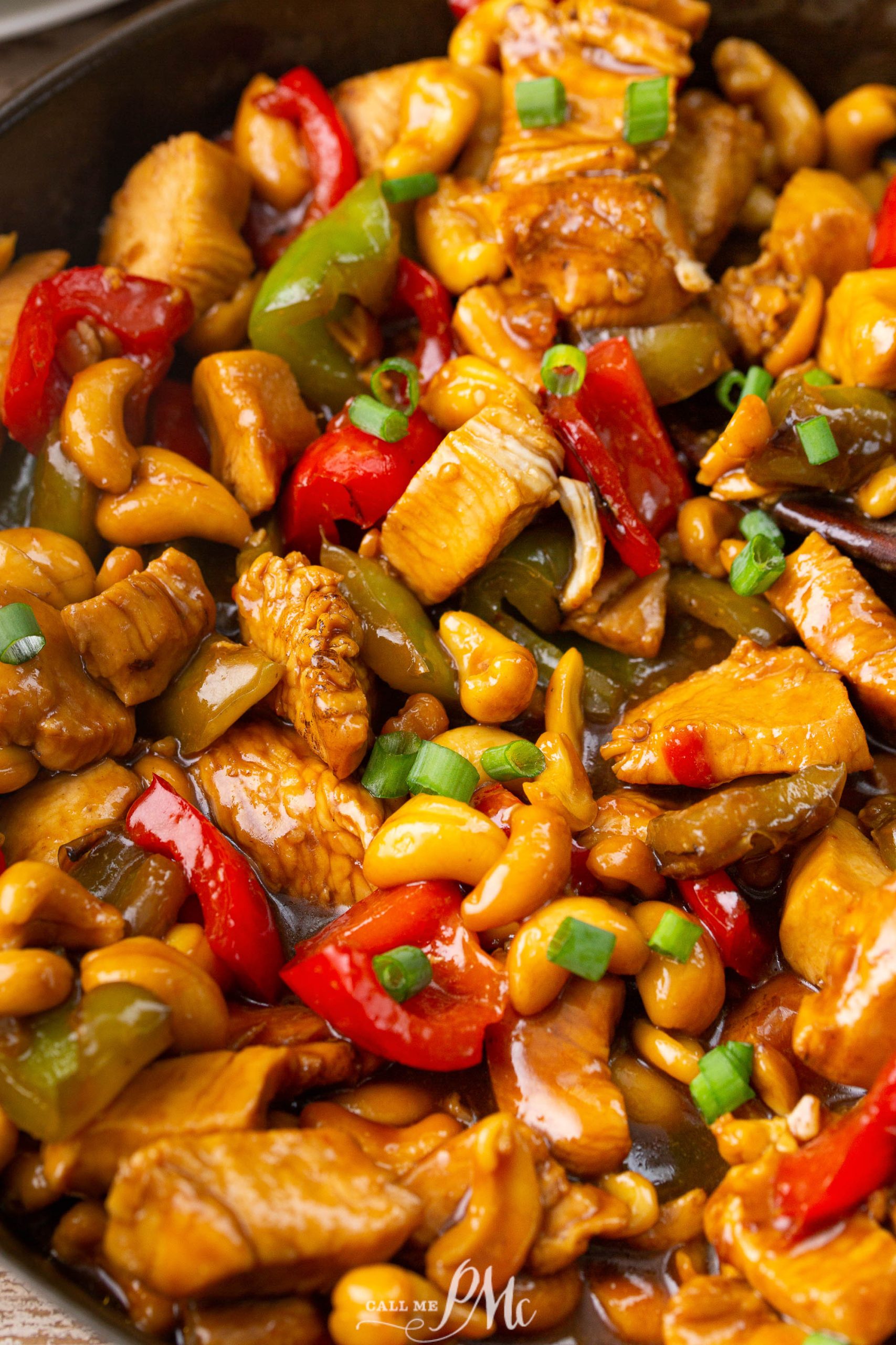 Close-up of a stir-fry dish with chicken, bell peppers, cashews, and green onions in a glossy sauce.