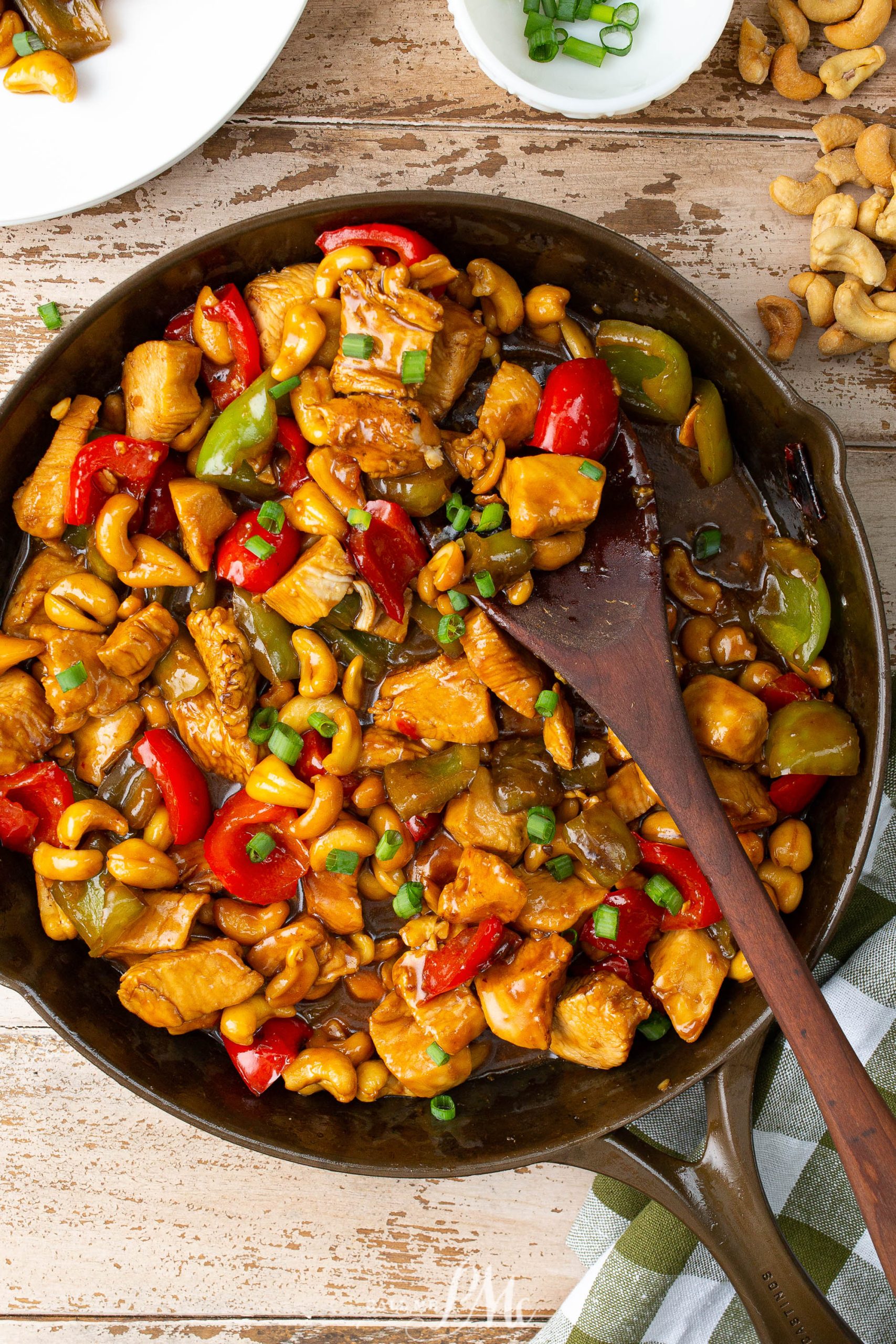 A skillet filled with cashew chicken stir-fry, featuring diced chicken, bell peppers, cashews, and green onions in a savory sauce, beside a white plate and scattered cashews.