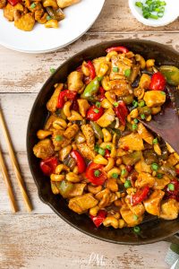 A skillet filled with cashew chicken stir-fry, featuring diced chicken, colorful bell peppers, and cashews, garnished with green onions. Chopsticks and a plate with a portion of the dish are on the side.