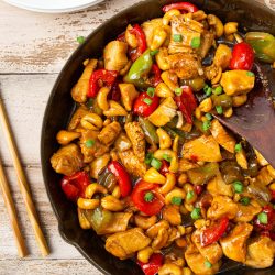 A skillet filled with cashew chicken stir-fry, featuring diced chicken, colorful bell peppers, and cashews, garnished with green onions. Chopsticks and a plate with a portion of the dish are on the side.