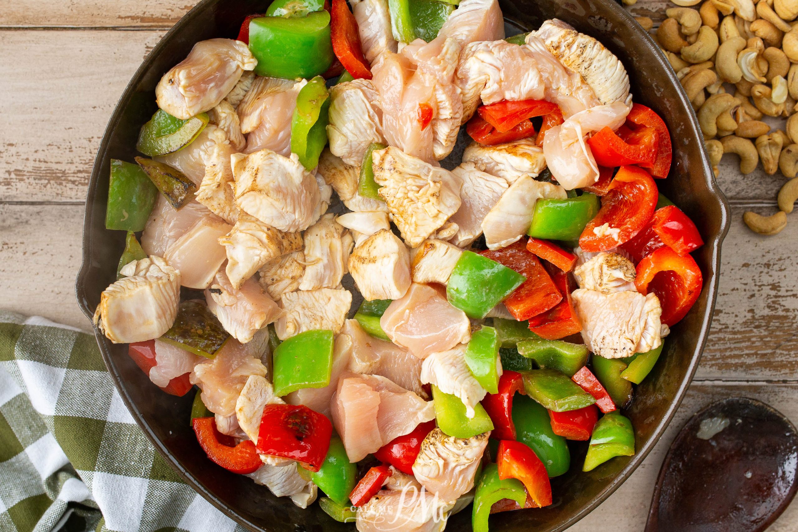 A skillet filled with uncooked chicken pieces, chopped green and red bell peppers. A green and white checkered cloth and some cashews are placed nearby.