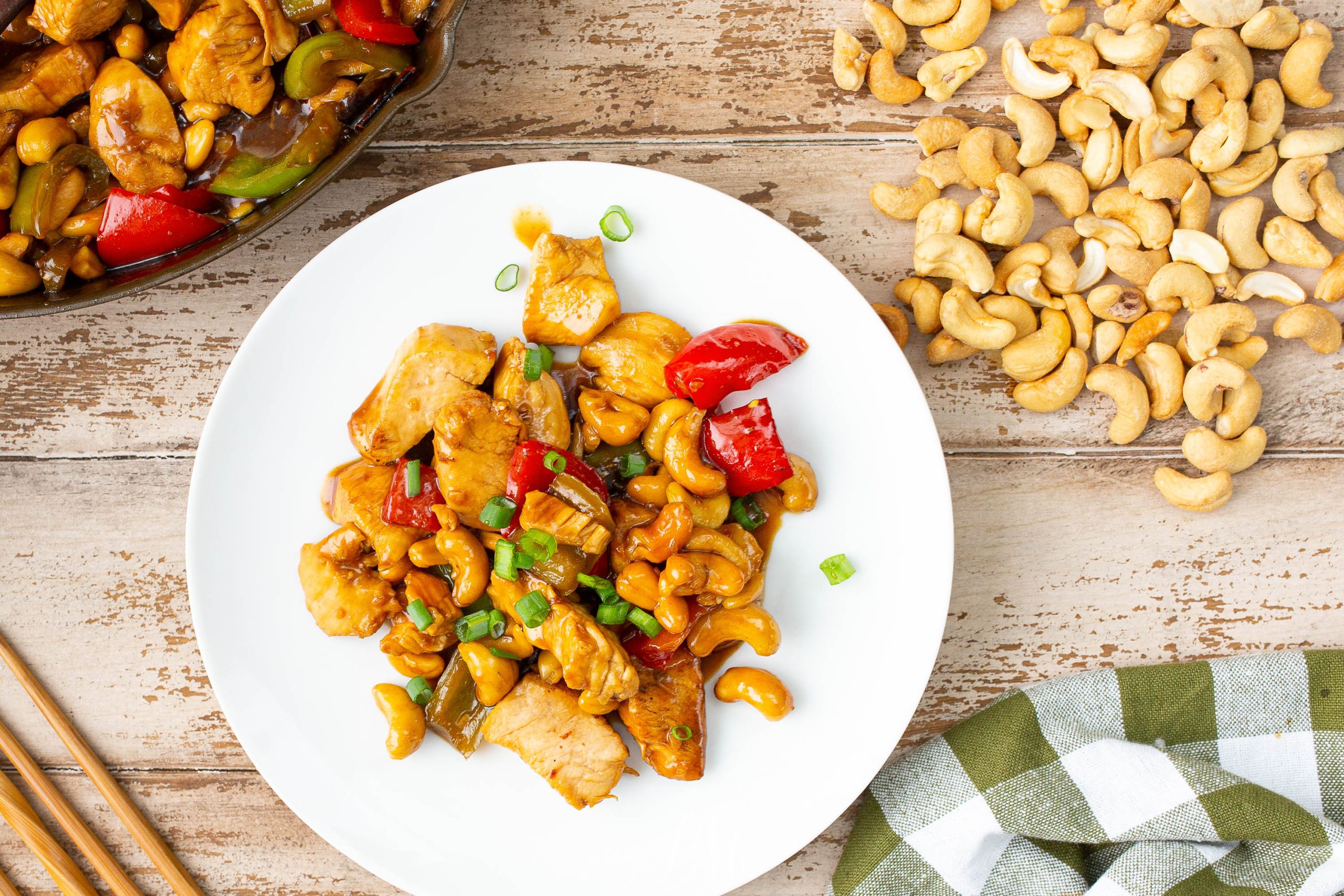 A plate of chicken with cashew nuts, bell peppers, and green onions is placed on a wooden surface next to a cast iron skillet containing more of the dish. A handful of cashews and a checkered napkin are beside the plate.