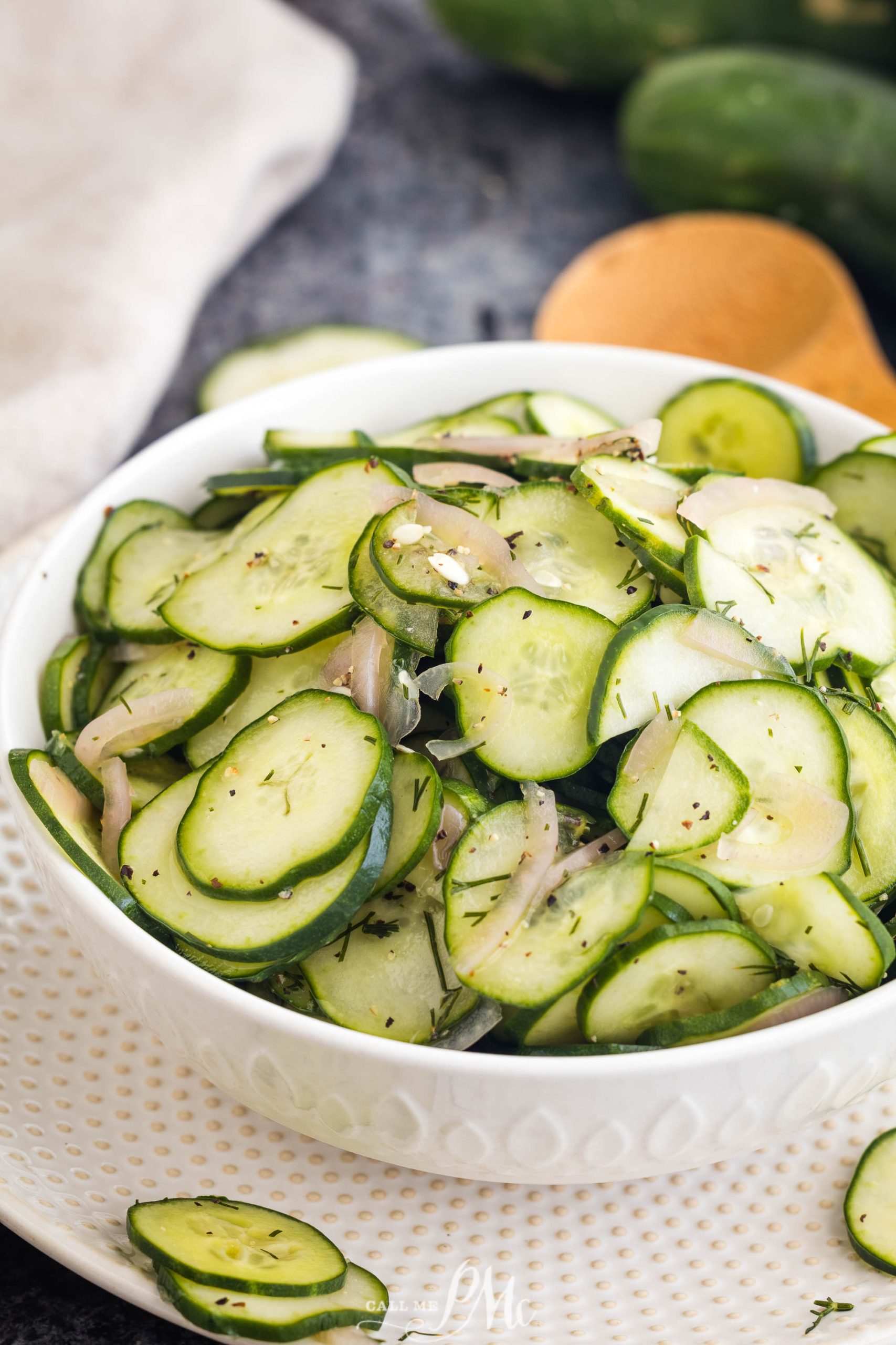 A white bowl filled with sliced cucumbers and onions, garnished with dill and black pepper, placed on a textured white plate. Cucumbers and a napkin are in the background.