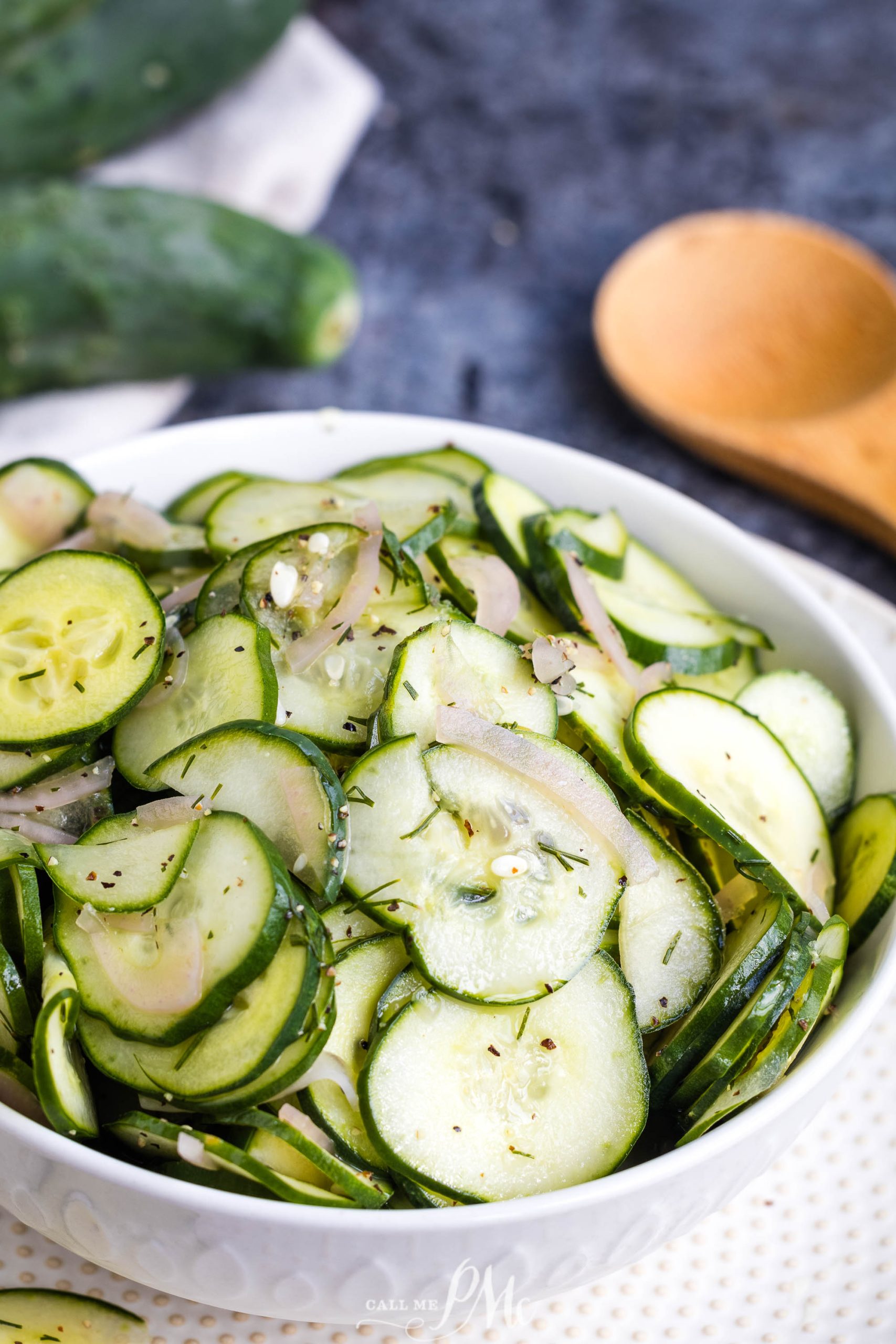 A white bowl filled with sliced cucumbers, onions, and herbs, with a wooden spoon and whole cucumbers in the background.