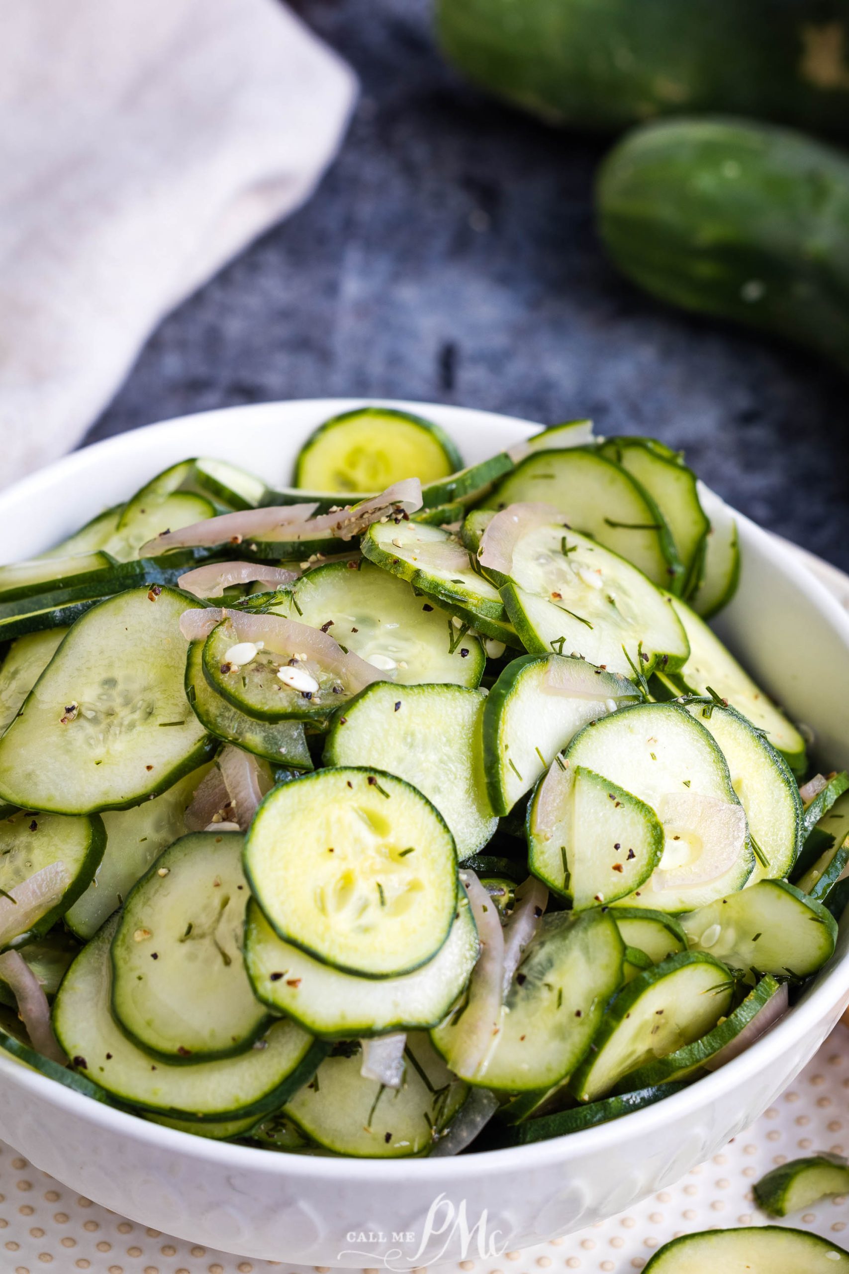 A bowl filled with thinly sliced cucumbers, onions, and herbs, set on a white surface with whole cucumbers in the background.